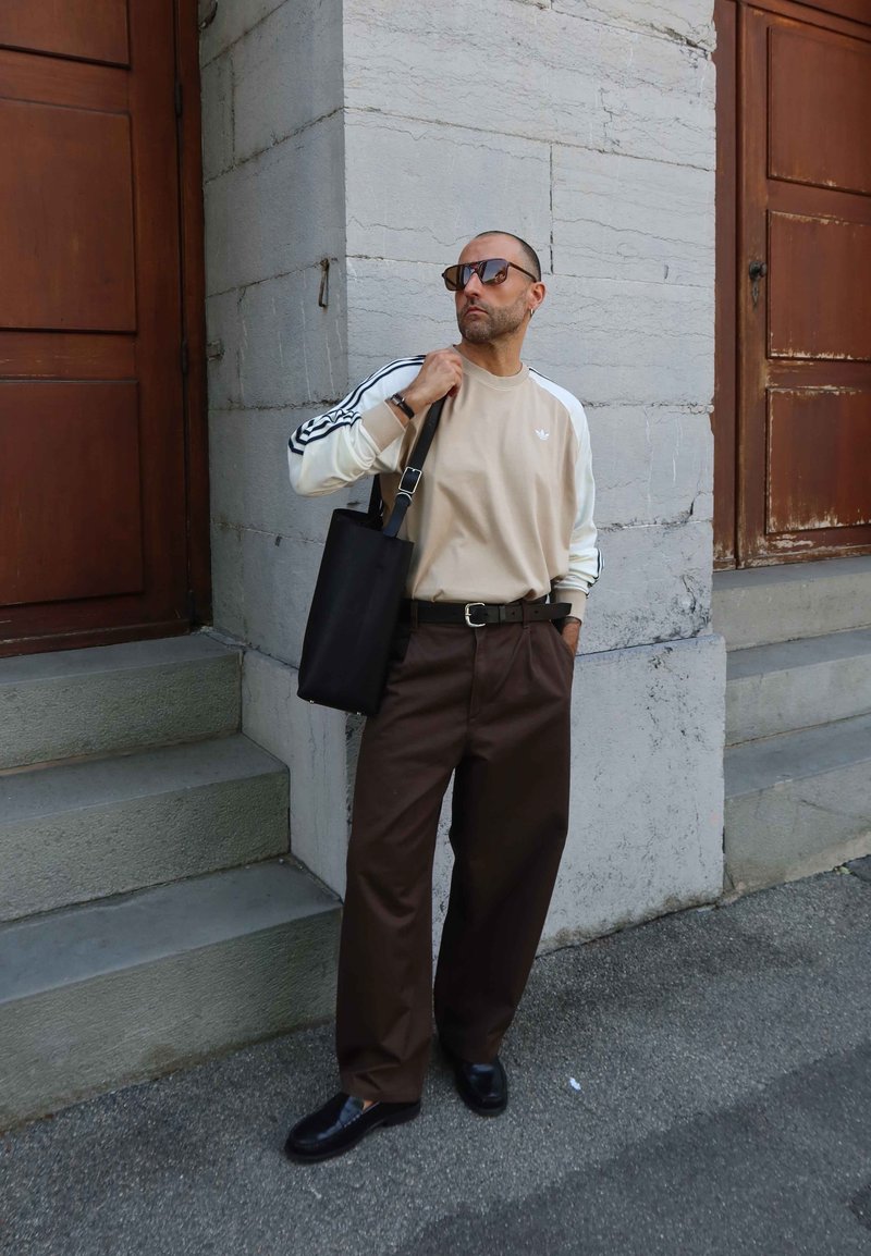 Homme portant des lunettes de soleil, une chemise beige et blanche à manches longues, un pantalon brun foncé, des chaussures noires et portant un sac fourre-tout noir, debout au coin en pierre d’un bâtiment.