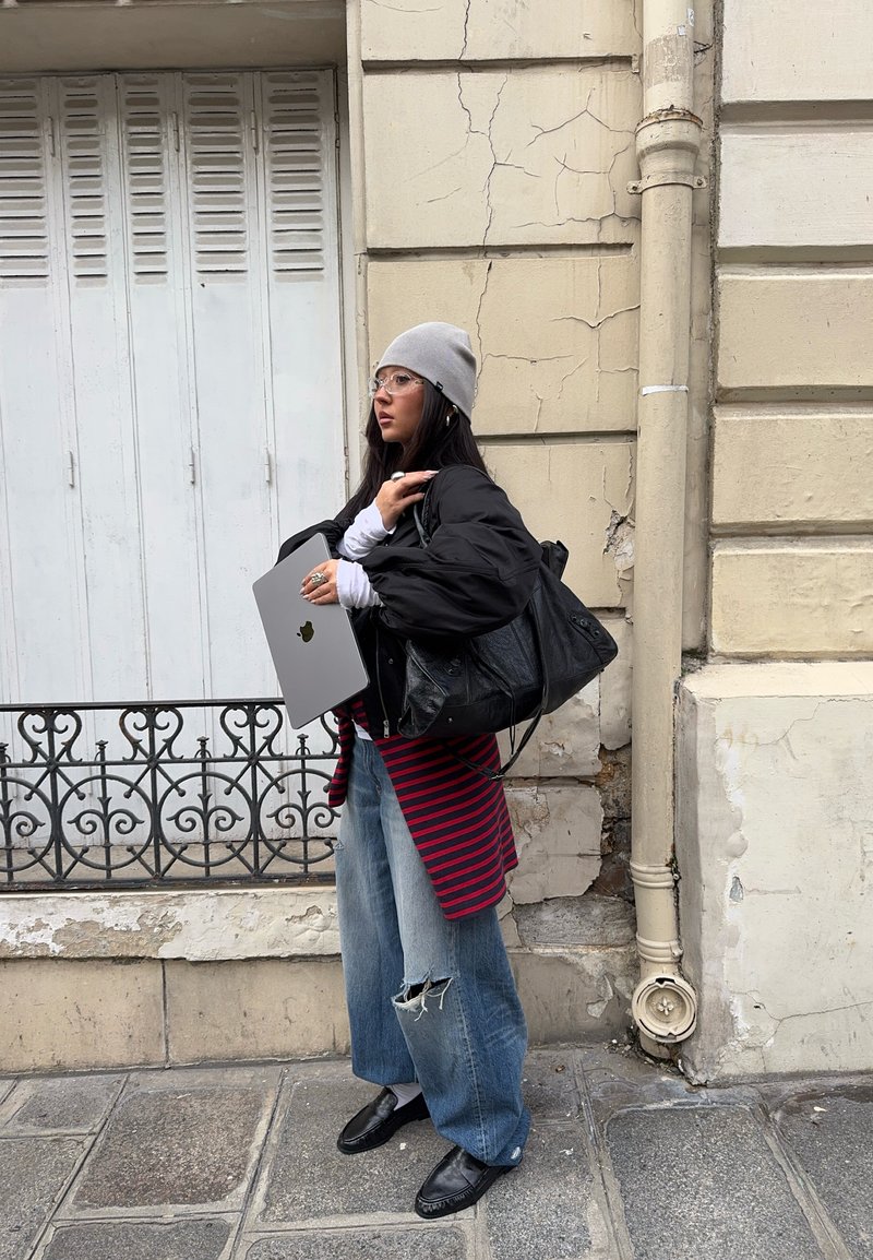 Tânără în haine casual ținând un laptop și o geantă mare neagră, stând pe trotuarul unui oraș lângă un zid bej uzat.