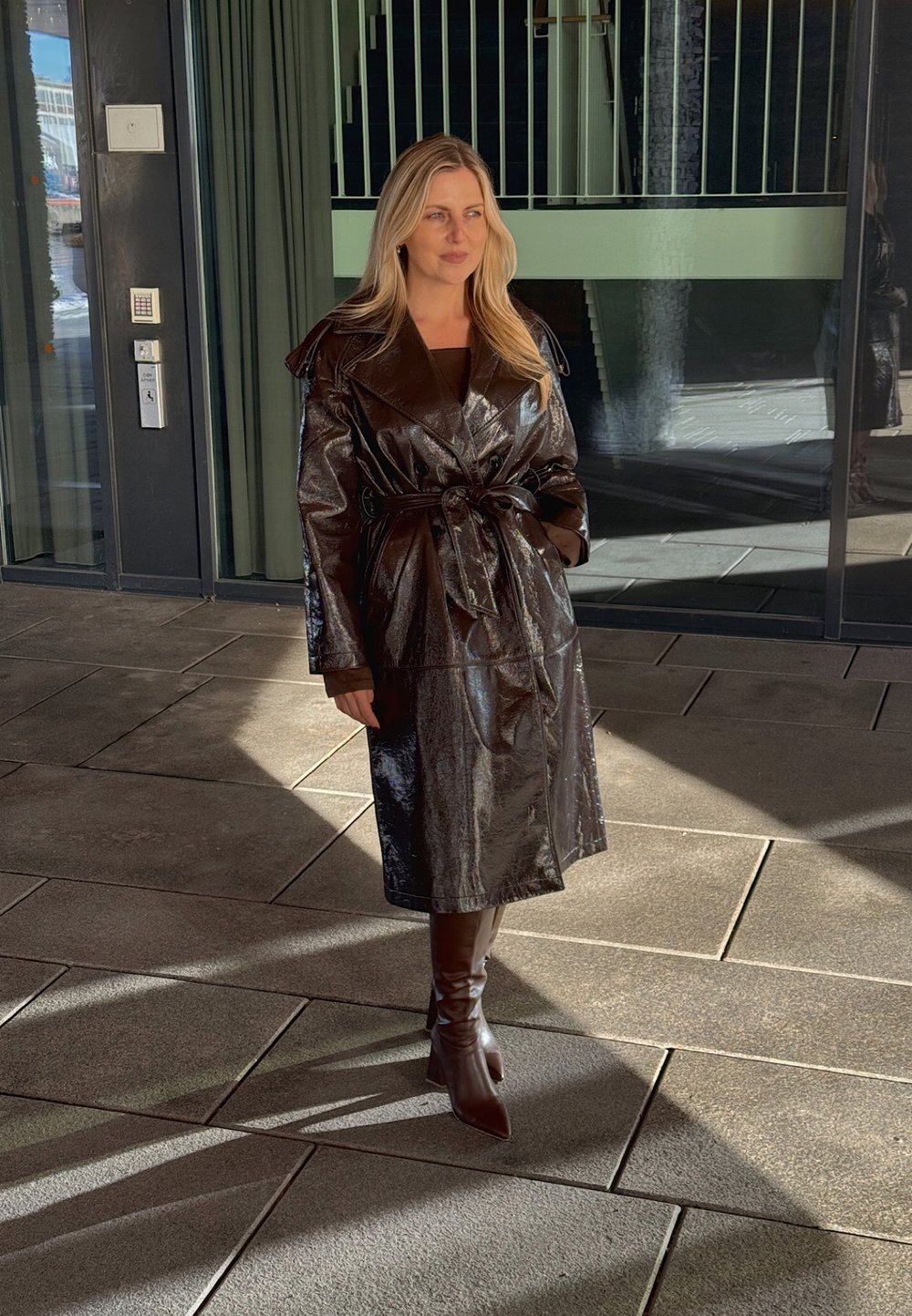 Brauner, glatter, langer Trenchcoat mit übergroßen Revers, tailliertem Gürtel und schmalen Ärmeln. Trägt dazu passgenaue braune Stiefel bis zum Knie.