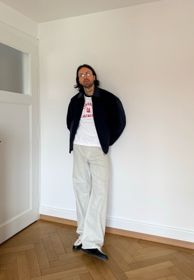 Hombre con gafas y tatuajes apoyado en una pared blanca, vestido con una chaqueta negra, camiseta blanca con estampado, pantalones anchos blancos y zapatos blanco y negro.