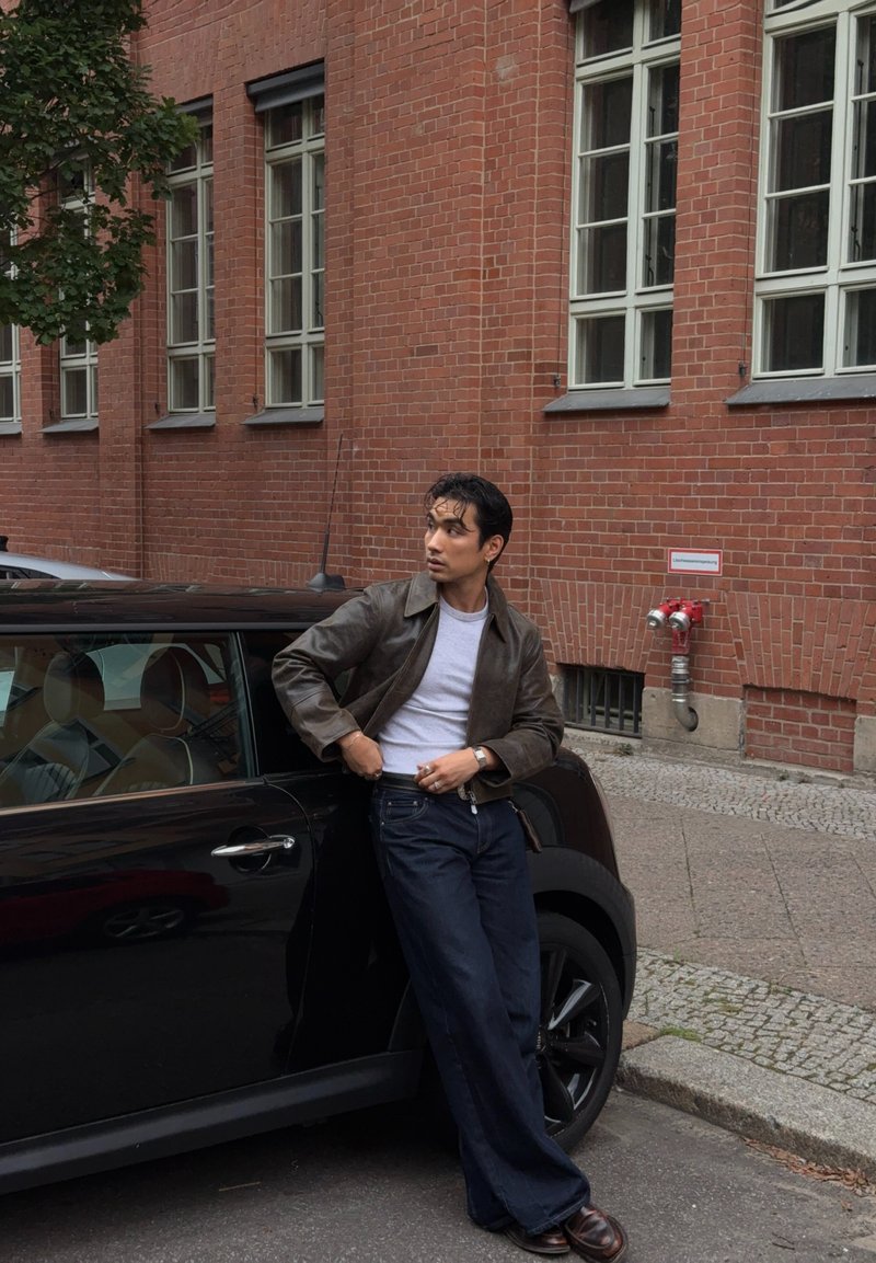 Homem a usar uma jaqueta castanha, camisa branca e jeans escuros encostado a um carro preto numa rua da cidade com edifícios de tijolo vermelho.