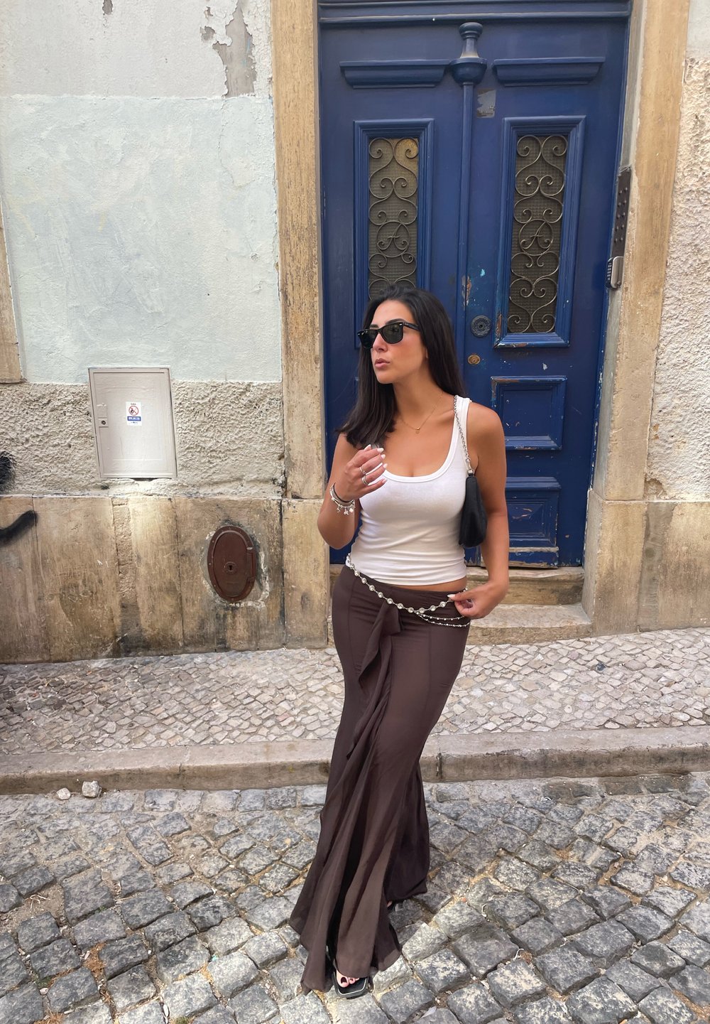 Brown, high-waisted, flowing trousers with a tied waist detail, paired with a fitted white tank top. Black sunglasses and a shoulder bag complement the outfit.
