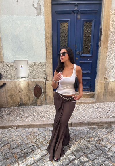 Pantalones fluidos marrones de cintura alta con detalle de cintura atada, combinados con una camiseta sin mangas blanca ajustada. Gafas de sol negras y un bolso de hombro complementan el look.