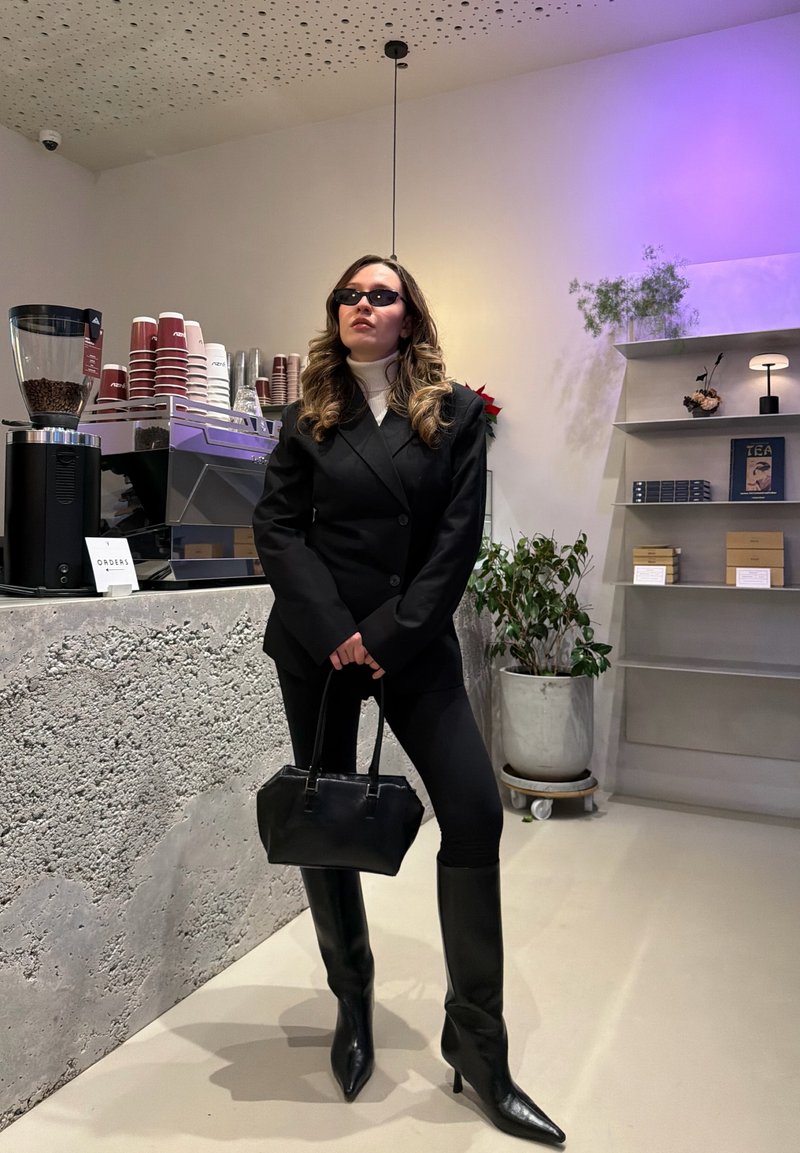 Blazer noir croisé porté sur un col roulé blanc, pantalon noir ajusté, bottes pointues montant jusqu'aux genoux, et petit sac à main noir.