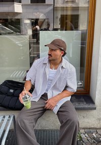 Pruhovaná krátká košile s krátkým rukávem, bílá a modrá, přes bílý top, zkombinovaná s širokými hnědými kalhotami; drží průhledný hrnek se zelenou tekutinou.
