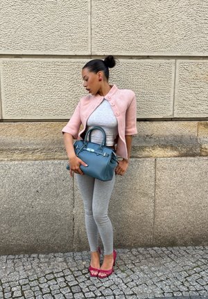 Mujer con cárdigan rosa y leggings grises sosteniendo un bolso azul, parada frente a una pared de piedra texturizada sobre un pavimento adoquinado.