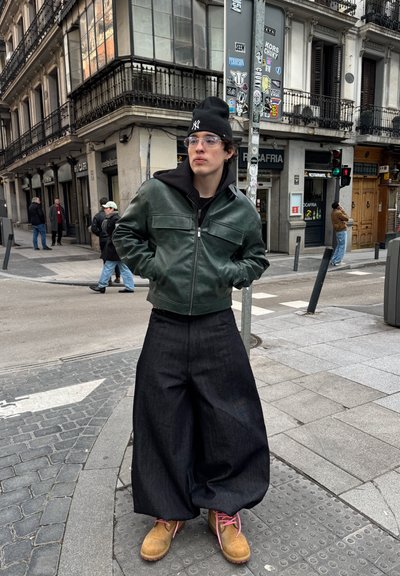 Persona joven con gorro negro, chaqueta de cuero verde, pantalones oscuros holgados y botas color canela, de pie en una esquina urbana.