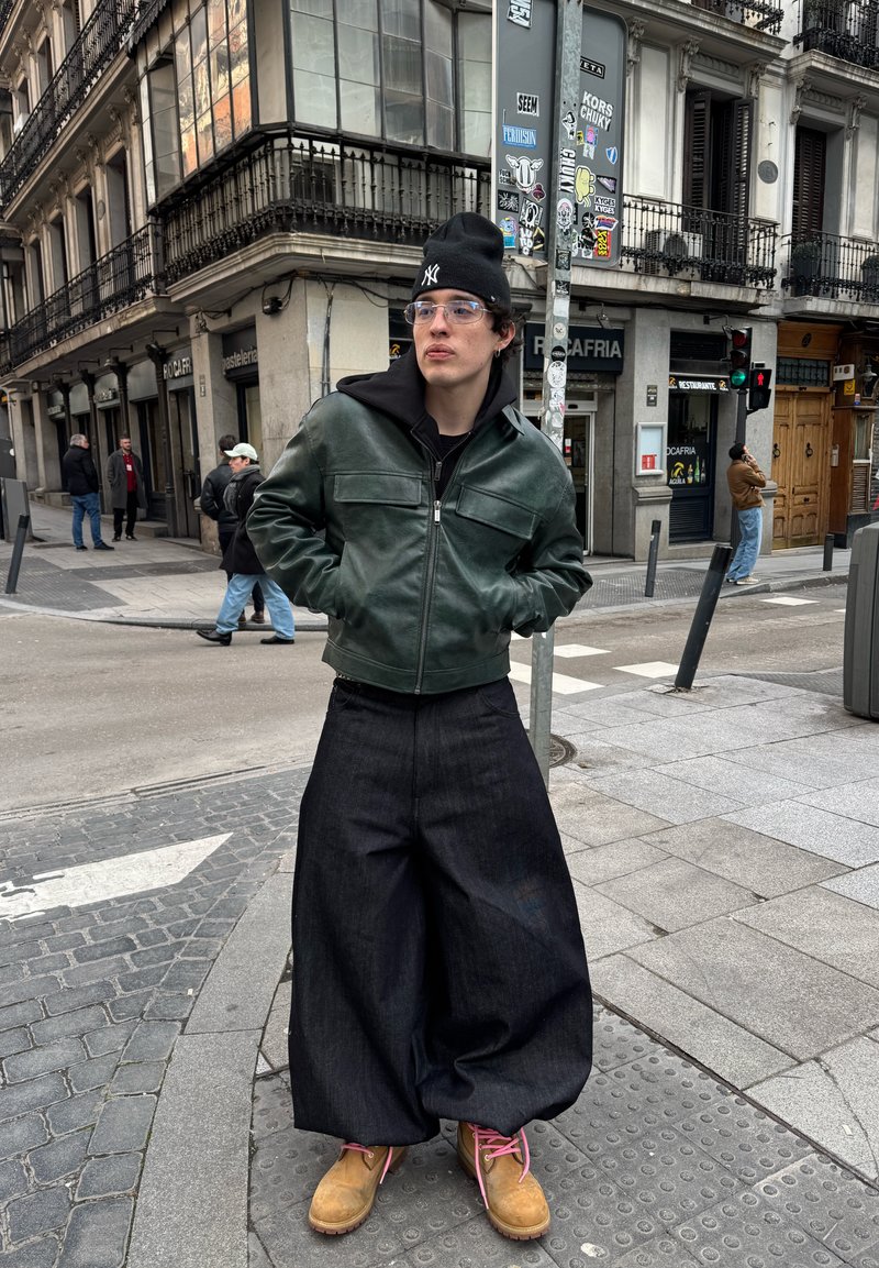 Jeune personne portant un bonnet noir, une veste en cuir verte, un pantalon large sombre et des bottes beige debout à un coin de rue urbain.