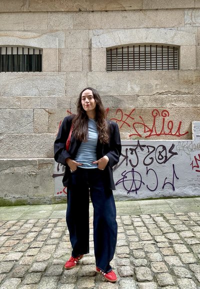 Mujer de pie en una calle adoquinada, vestida con chaqueta negra, camisa azul, vaqueros anchos y zapatos rojos, con grafitis en una pared de piedra tras ella.