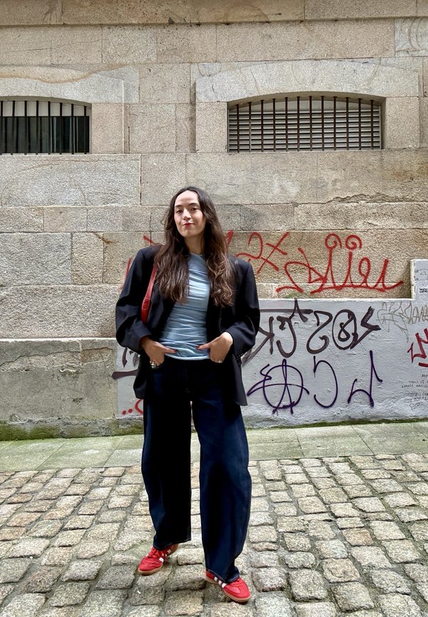 Žena stojí na dláždenej ulici, má na sebe čiernu bundu, modrú košeľu, široké rifle a červené topánky, za ňou je na kamennom múre graffiti.