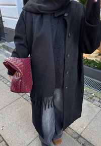 Black wool coat with button fastening and fringed hem, layered over a grey jumper. Holding a burgundy woven clutch. Grey jeans, brown boots.