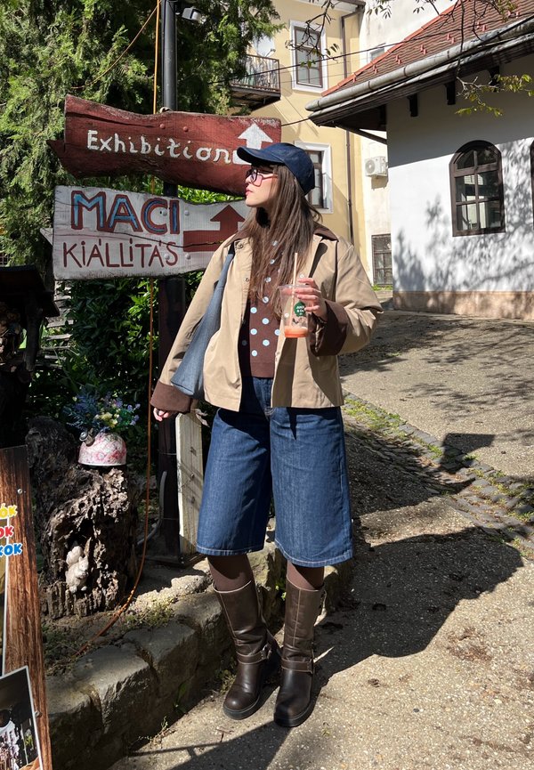 Frau in Freizeitkleidung hält ein Getränk und steht nahe bei hölzernen Schildern, die auf eine Ausstellung und ein Museum an einer sonnigen Straße zeigen.