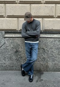 Man med grå tröja, blå jeans, svarta skor och brun keps som lutar sig mot en texturerad stenvägg med armarna i kors.