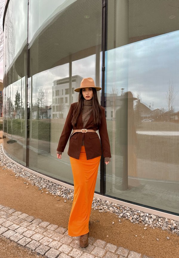 Brauner Blazer mit einem Gürtel über einem braunen Rollkragenpullover, kombiniert mit einem langen orangefarbenen Rock und einem beigen Hut, der in der Nähe eines Gläsernen Gebäudes steht.