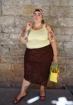 Persona con tatuajes que lleva una camiseta sin mangas amarilla, una falda de encaje marrón, un pañuelo en la cabeza y sandalias, sosteniendo un pequeño bolso tejido amarillo, de pie junto a una pared de piedra.