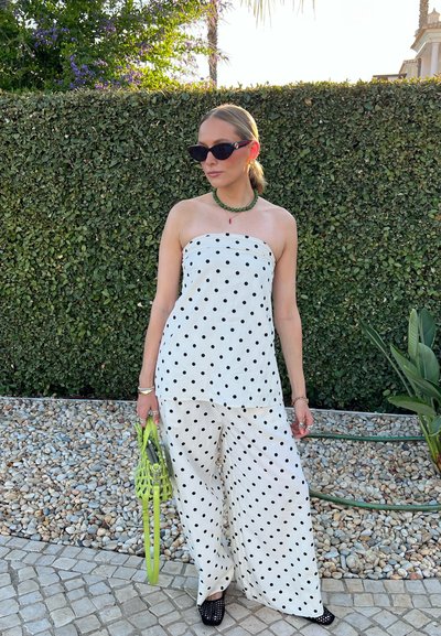 Conjunto blanco sin tirantes con lunares negros, pantalones de pierna ancha, sosteniendo un bolso de mano tejido verde brillante, con un fondo de vegetación texturizada.