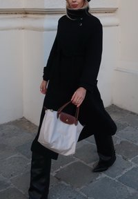 Černý kabát s pásem v pase, doplněný lesklými černými legínami. Světle zbarvená kabelka s hnědými akcenty, na pozadí kamenné dlažby.