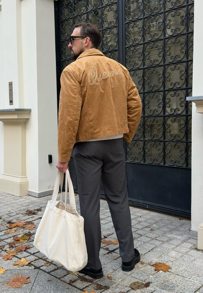 Hombre con chaqueta de ante color beige con "Queens" bordado en la parte trasera, pantalones grises, zapatos negros y gafas de sol, sostiene una gran bolsa de tote blanca en una calle empedrada.