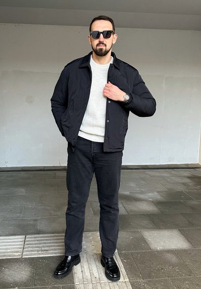 Chaqueta de plumas negra sobre un suéter claro, combinada con jeans negros y zapatos negros brillantes. El fondo presenta un espacio de hormigón con suelo de baldosas.