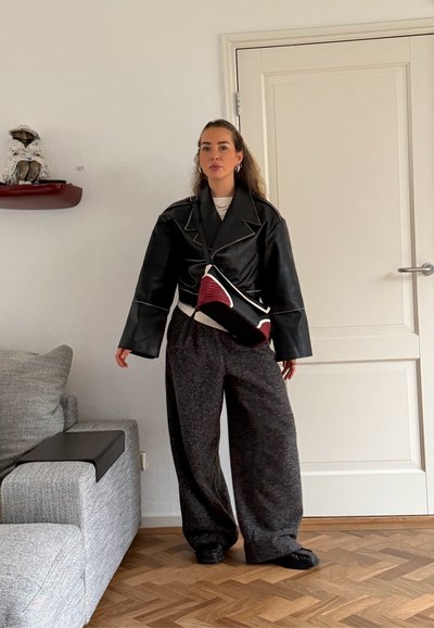 Mujer de pie en interiores, vistiendo una chaqueta de cuero negra sobredimensionada, pantalones anchos grises, zapatos negros y un bolso bandolera negro con rojo.