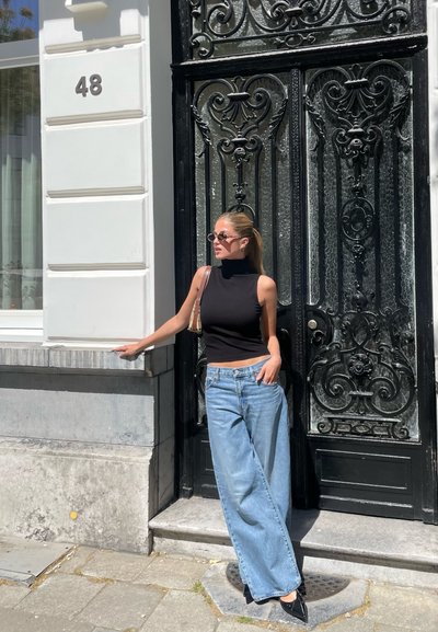 Top negro sin mangas con cuello alto, jeans de pierna ancha en azul claro y zapatos de tacón negro, con un fondo de una puerta negra ornamentada y detallada.