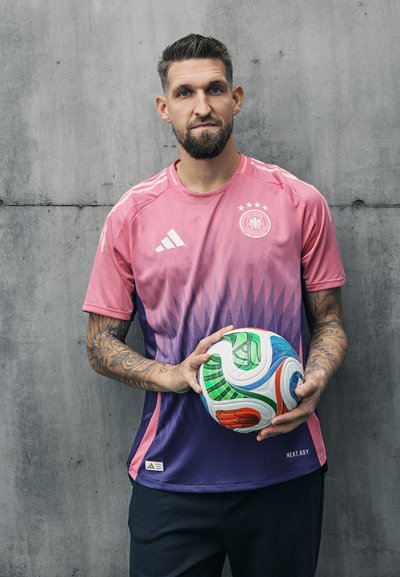 Camisa de fútbol en degradado rosa y púrpura con tres estrellas y el logo de Adidas. El modelo sostiene un balón de fútbol multicolor. Fondo de concreto.