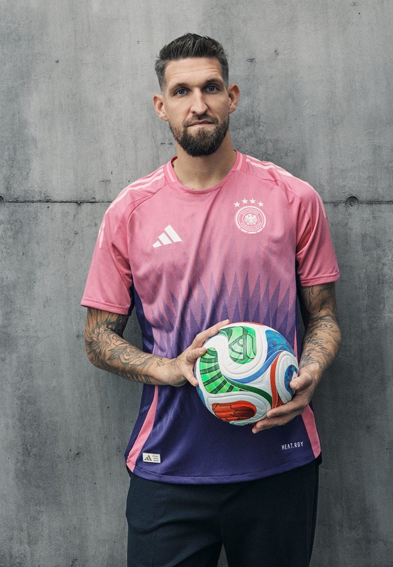 Maillot de football dégradé rose et violet avec trois étoiles et logo Adidas. Le modèle tient un ballon de football multicolore. Fond en béton.