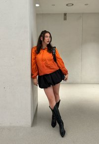 Oranžová bunda na zips s nafúknutými rukávmi, čierna plisovaná sukňa a čierne čižmy po kolená. Doplnok: čierny ruksak s cvokmi. Hladké, neutrálne pozadie.