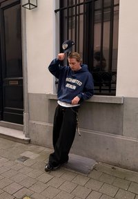 Jeune homme appuyé sur un rebord de bâtiment, tenant une casquette au-dessus de sa tête, portant un sweat à capuche bleu marine avec le texte « PROHIBITED BERLIN » et un pantalon noir ample.