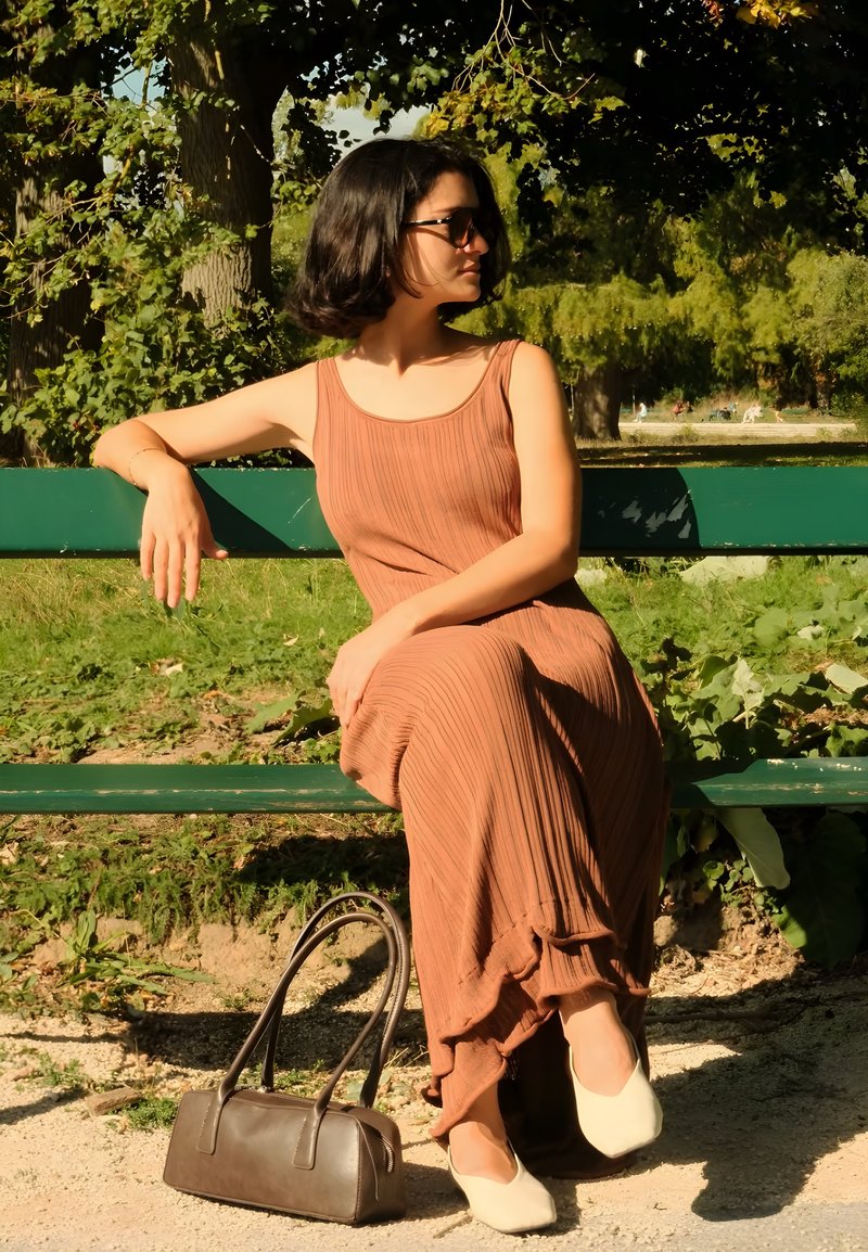 Maxi-Kleid in Braun mit geripptem Muster und rüschendem Saum, kombiniert mit hellen Ballerinas und einer braunen Handtasche. Sitzt auf einer grünen Bank in einer Parkumgebung.