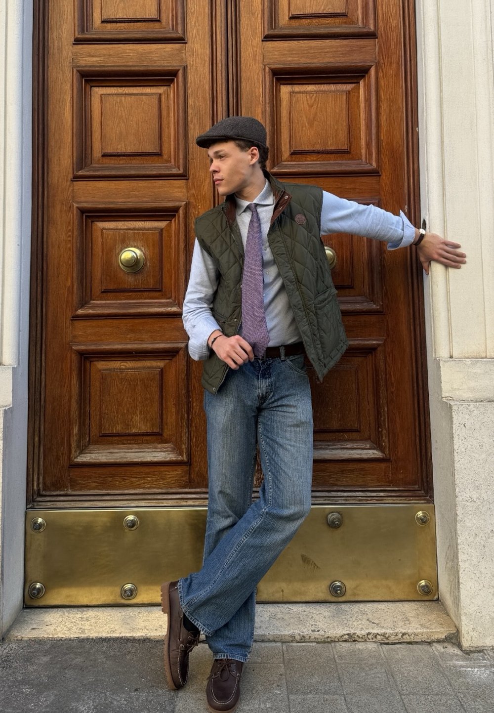 Gilet trapuntato verde, camicia azzurro chiaro, cravatta viola, jeans blu, scarpe marroni, berretto piatto; appoggiato a una porta di legno con maniglia circolare in ottone.