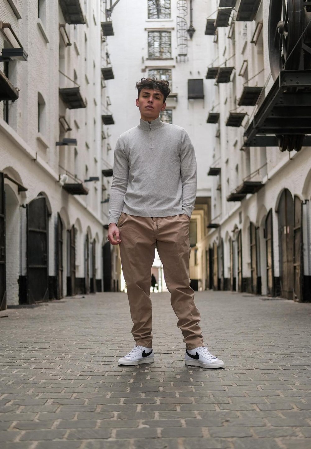 Light grey zip-neck sweater, tapered tan trousers, and white trainers with black accents, standing on cobblestones in an alleyway with brick walls.