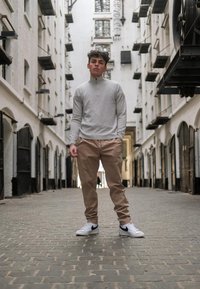 Light grey zip-neck sweater, tapered tan trousers, and white trainers with black accents, standing on cobblestones in an alleyway with brick walls.