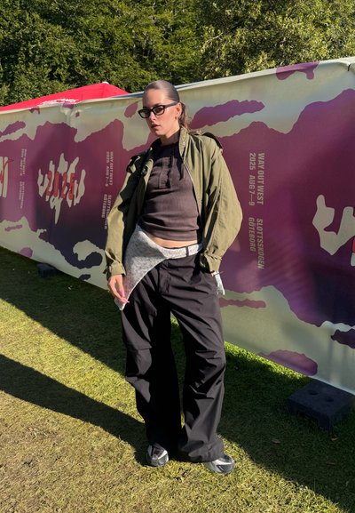 Chaqueta bomber verde, top marrón ajustado, pantalones grises con cintura blanca, pantalones negros oversize, zapatillas grises y gafas de sol frente a un banner colorido.