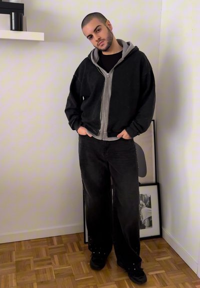 Sudadera negra con cremallera sobre una camiseta gris, combinada con unos pantalones negros sueltos de corte recto. La habitación tiene suelos de madera y fotos enmarcadas.