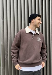 Sweatshirt castanha com gola cor creme, com texto bordado. Usada por cima de uma camisa branca, combinada com jeans cinzentos. Gorro preto na cabeça.