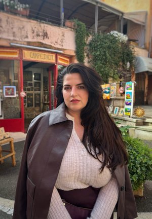 Abrigo de cuero marrón sobre un suéter de canalé claro, de pie frente a una vitrina colorida con plantas verdes y varias obras de arte.