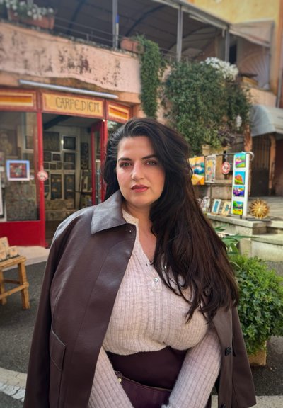 Abrigo de cuero marrón sobre un suéter de canalé claro, de pie frente a una vitrina colorida con plantas verdes y varias obras de arte.