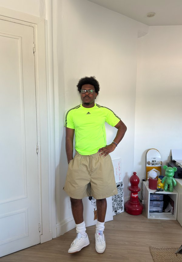 Man wearing neon green Adidas shirt and beige shorts stands in a room with skateboard, large red chess piece, and green figurine on shelves.