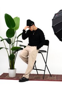 Pulover negru texturat, pantaloni bej, pantofi negri. Așezat pe un scaun lângă o plantă mare verde. Lumină difuză provenind de la o umbrelă adiacentă.