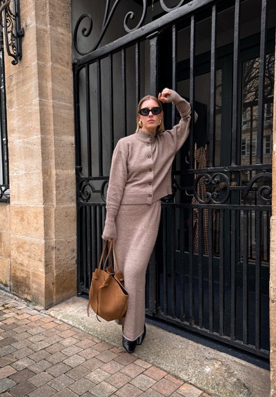 Suéter de punto beige con botones y falda larga a juego. Botas negras de tacón alto, bolso grande marrón y gafas de sol. Entorno al aire libre.