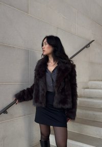 Donna che indossa un lungo cappotto di pelliccia scura, una gonna nera e stivali, in piedi su una scala di marmo, mentre si tiene al corrimano e guarda verso sinistra.