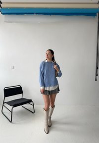 Světle modrý pletený svetr přes bílou košilovou halenku, černobílé skládací sukně a béžové kovbojské boty. V pozadí černá židle.