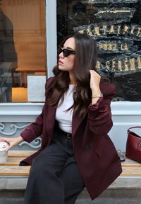 Blazer color bordeaux sopra una camicia bianca, abbinato a pantaloni neri a gamba larga; tiene in mano una tazza di caffè, una borsa rossa visibile nelle vicinanze. Occhiali da sole neri completano il look.