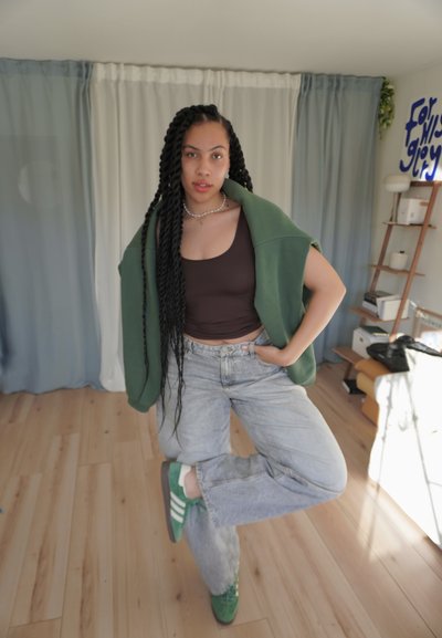 Cardigan verde de gran tamaño, top corto marrón, vaqueros baggy azul claro y zapatillas verdes con rayas blancas. Pelo largo trenzado con un collar.