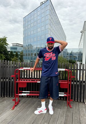 Jersey de marina con acentos rojos y numeración "05", combinado con pantalones cortos de mezclilla oscuros y zapatillas blancas con rayas rojas. Barandilla de metal en el fondo.