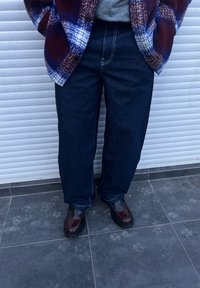 Temno modri džins hlače s ravnim krojem, poudarjene z svetlimi šivi, v kombinaciji z sijočimi rjavimi loaferji in karirasto večbarvno jakno.