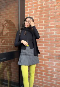 Femme aux longs cheveux noirs portant un pull noir, une jupe grise à fines rayures et des collants jaune vif, debout contre un mur de briques, main sur le front.