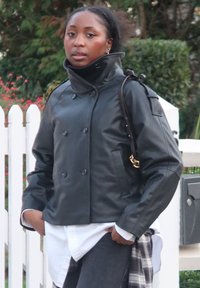 Svart dubbelknäppt skinnjacka med hög krage, detaljerad sömnad och axelrem. Bärs över en vit skjorta och mörka jeans i denim.