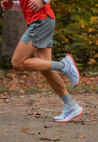 Kevyet juoksukengät, joissa on vaaleansininen päällinen, violetit yksityiskohdat ja punaiset pohjat. Puettuna harmaiden shortsien ja niihin sopivien sukkien kanssa juostessa polulla.